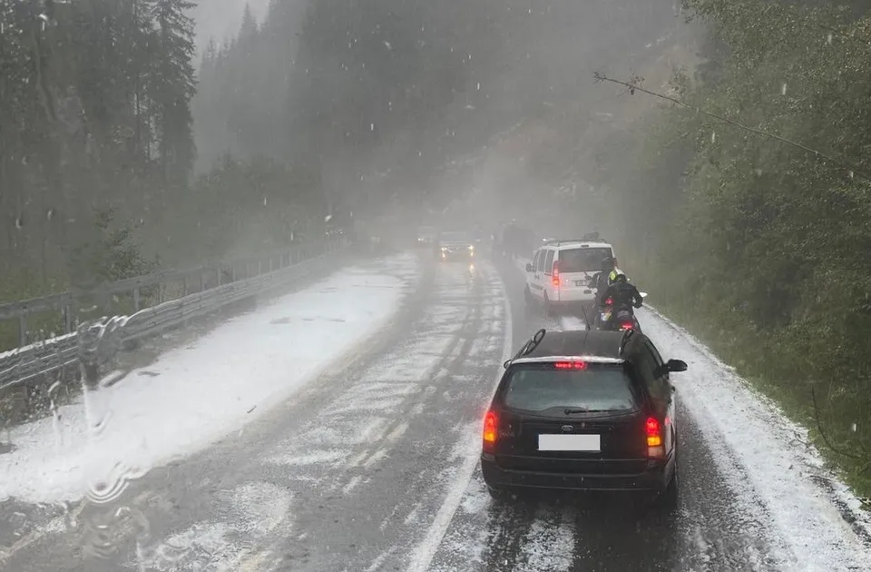 Mașini circulă pe un drum montan asfaltat, acoperit parțial de zăpadă, în condiții de vizibilitate redusă din cauza precipitațiilor și a ceții.
