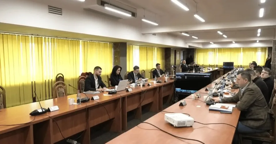Oamenii stau la mese lungi de conferință într-o sală cu jaluzele galbene, folosind laptopuri și microfoane pe suprafețele de lucru din lemn.