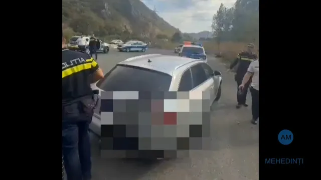O mașină albă stă oprită pe un drum, fiind înconjurată de mai multe persoane, inclusiv un agent de poliție cu un tricou negru și galben.