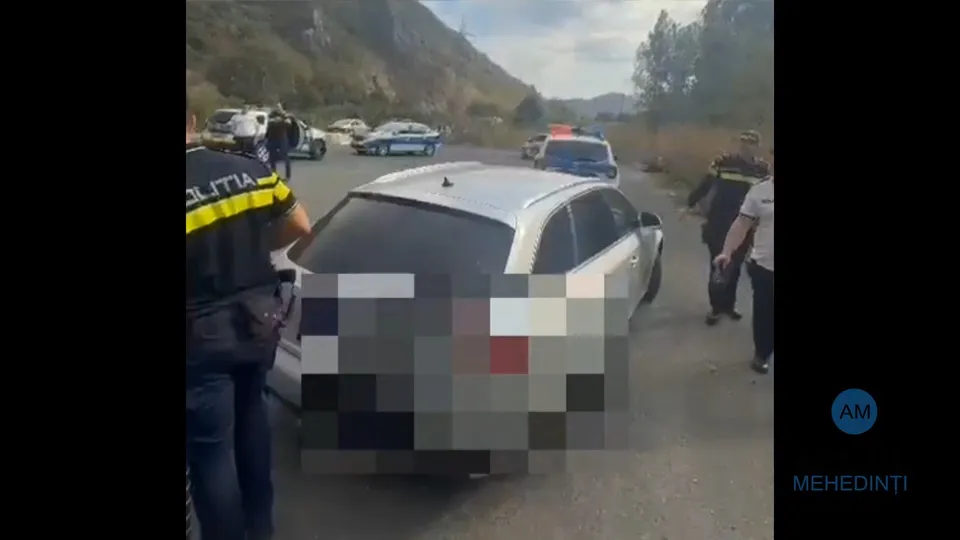 O mașină albă stă oprită pe un drum, fiind înconjurată de mai multe persoane, inclusiv un agent de poliție cu un tricou negru și galben.