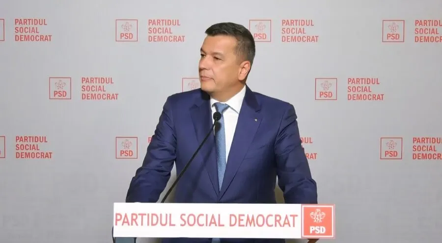 Un bărbat într-un costum albastru și cravată deschise, stând în fața unui fundal alb cu logourile roșii ale Partidului Social Democrat.
