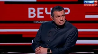 Un bărbat cu păr scurt și o jachetă închisă la culoare stă în fața unui fundal roșu și alb cu litere mari albe.