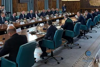Oamenii în costume purtând cămăși albe stau la o masă lungă de conferință într-o sală cu pereți din lemn.