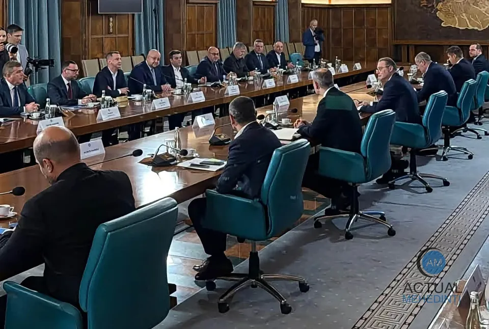 Oamenii în costume purtând cămăși albe stau la o masă lungă de conferință într-o sală cu pereți din lemn.