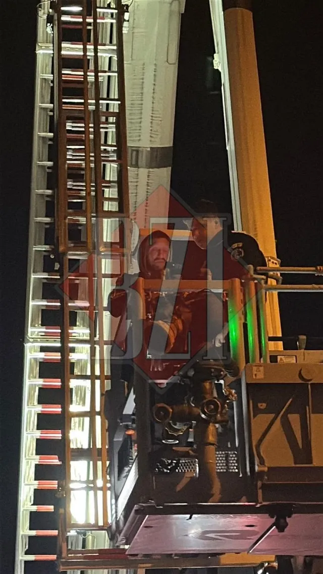 Două persoane stau pe o platformă ridicată a unei autospeciale de intervenție, lângă o scară metalică iluminată noaptea.
