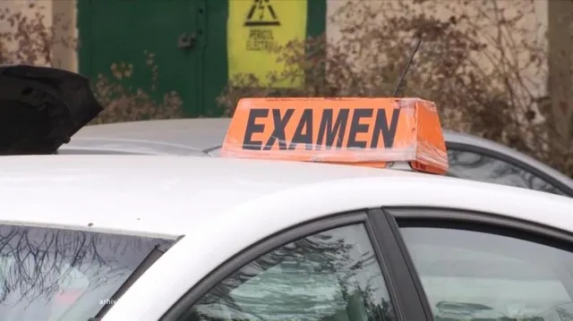 Un semn portocaliu cu textul negru EXAMEN este așezat pe acoperișul unei mașini albe lângă o ușă verde cu un avertisment galben.