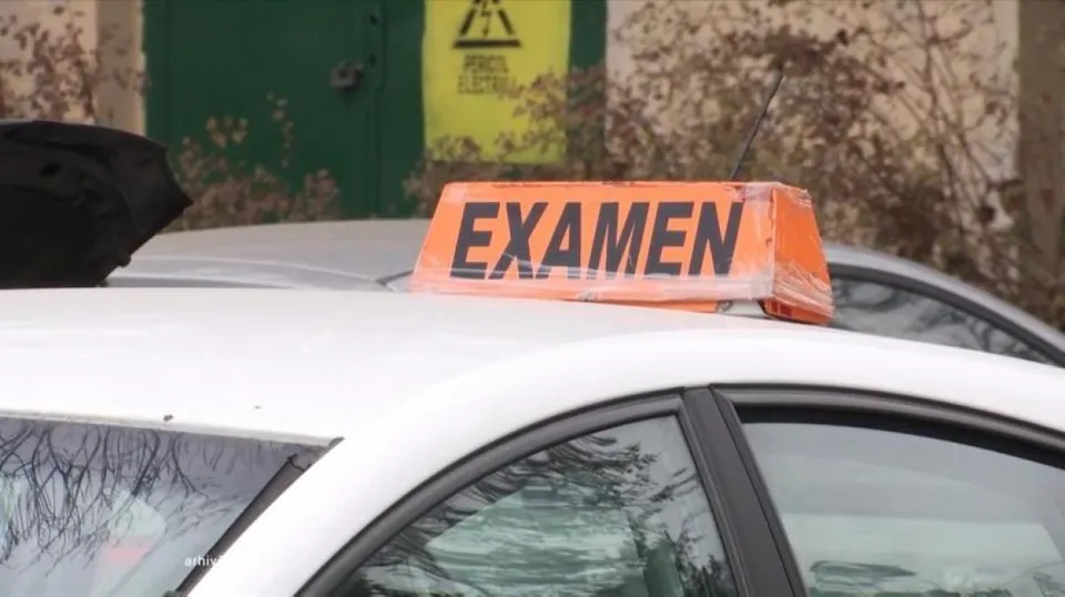 Un semn portocaliu cu textul negru EXAMEN este așezat pe acoperișul unei mașini albe lângă o ușă verde cu un avertisment galben.