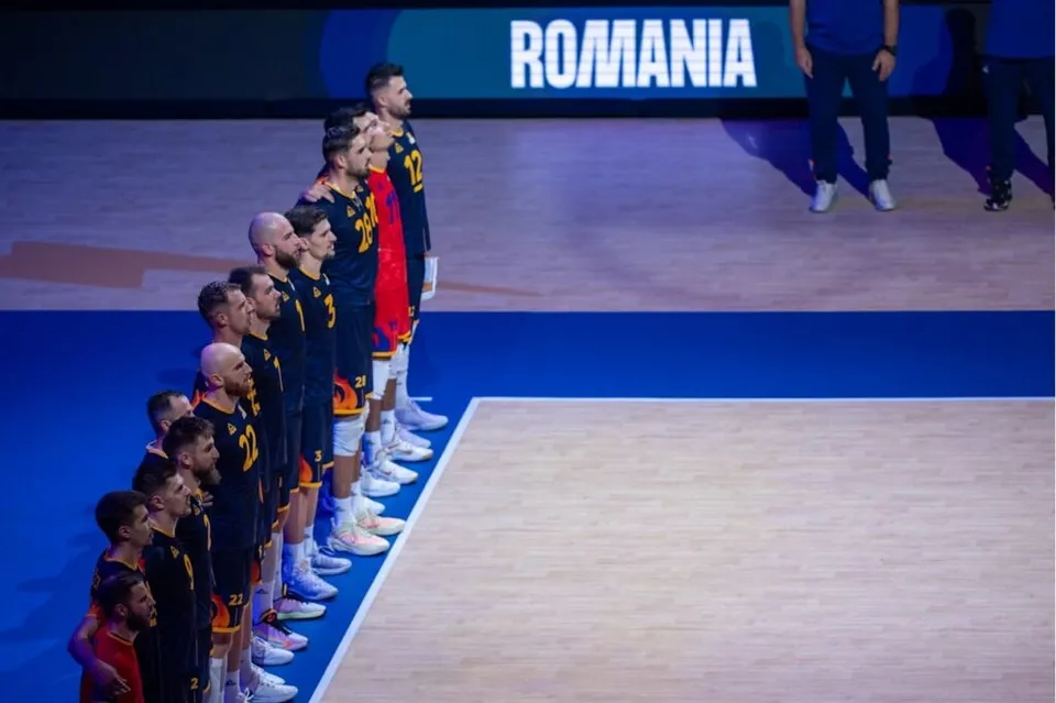 O linie de sportivi în echipamente de volei negre și roșii stă în picioare pe un teren de sport lângă un panou albastru.