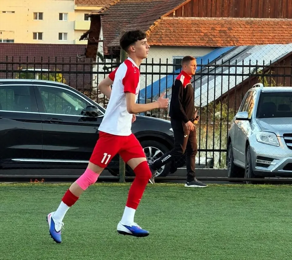 Un jucător de fotbal cu echipament alb și roșu aleargă pe un teren de iarba artificială, în timp ce un alt bărbat merge în fundal.