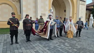 Oameni îmbrăcați în costume istorice stau pe o esplanadă pavată în fața unei clădiri cu arcade mari.