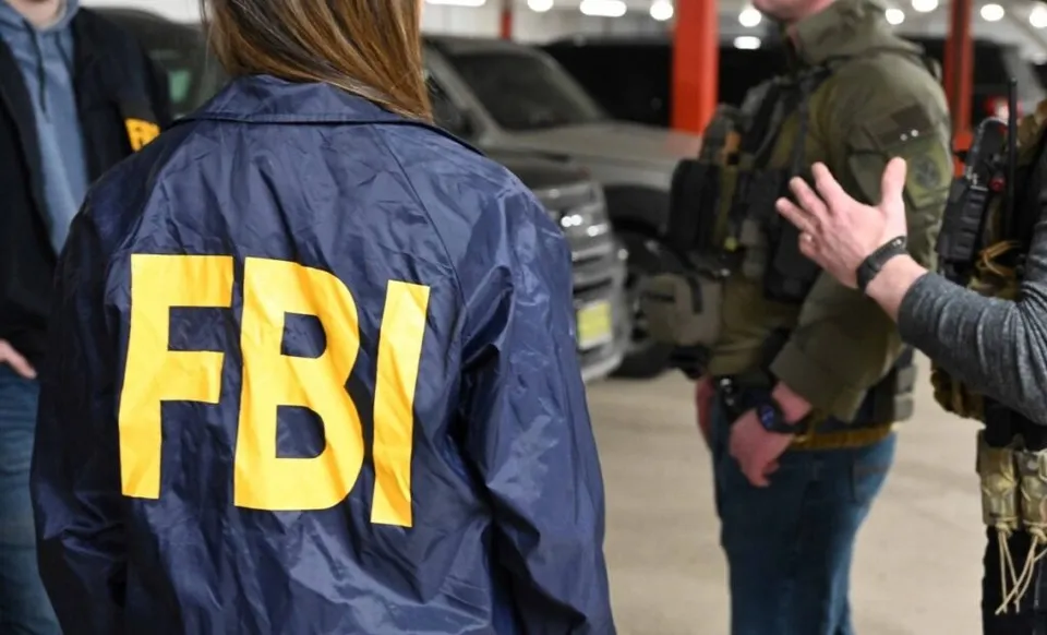 O persoană poartă o jachetă albastră cu inscripția FBI scrisă cu litere galbene, fiind întoarsă cu spatele față de cameră.