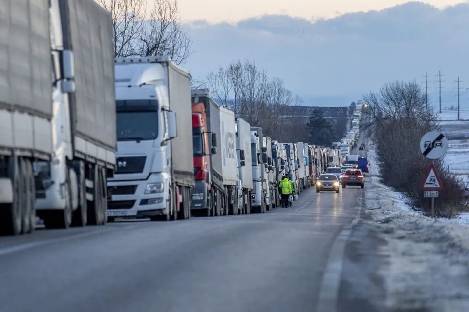 O coloană lungă de camioane mari este parcată pe marginea unui drum asfaltat într-un peisaj de iarnă cu zăpadă.