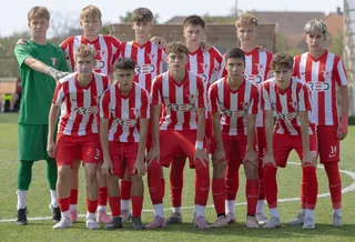 Un grup de adolescenți îmbrăcați în echipamente sportive roșii și albe pozează pe un teren de fotbal verde.