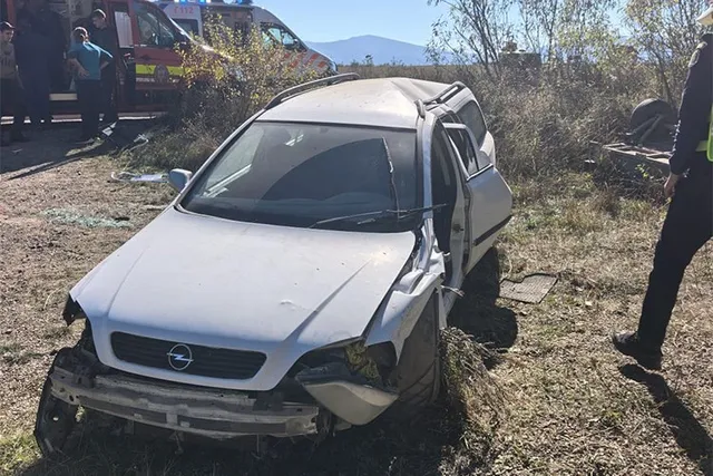 O mașină de culoare albă cu partea frontală și laterală stângă avariată stă într-un teren cu vegetație uscată lângă un vehicul de urgență.