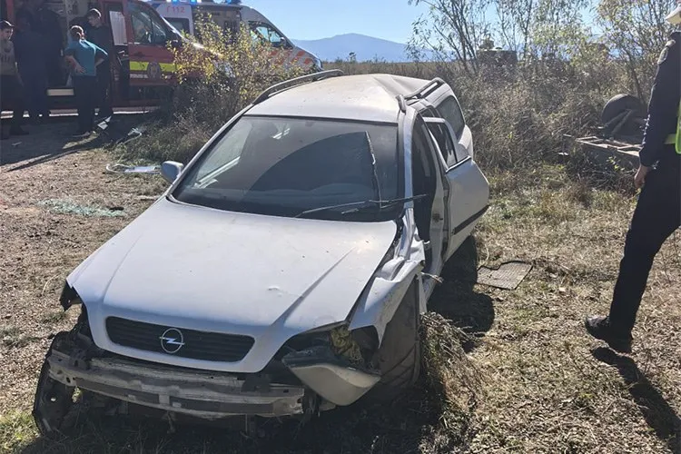 O mașină de culoare albă cu partea frontală și laterală stângă avariată stă într-un teren cu vegetație uscată lângă un vehicul de urgență.