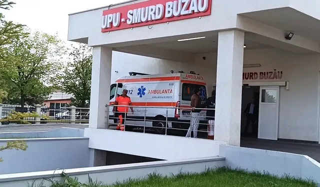 O ambulanță albă cu inscripții roșii și albastre este parcată sub un acoperiș de spital lângă un rampă de acces.