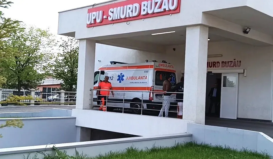 O ambulanță albă cu inscripții roșii și albastre este parcată sub un acoperiș de spital lângă un rampă de acces.