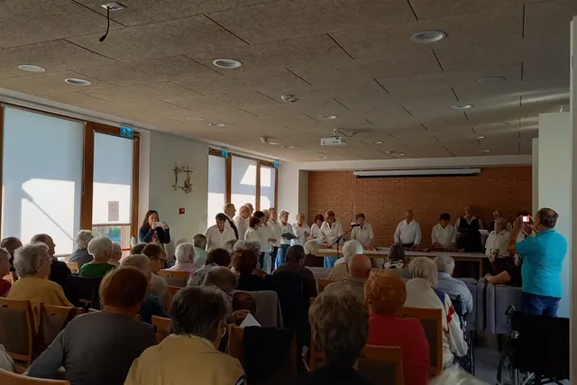 Un grup de persoane cântă într-o încăpere cu ferestre mari, în fața unui public așezat pe scaune.
