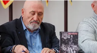 Un bărbat cu barbă căruntie și sacou închis peste o cămașă albastră stă la o masă lângă o carte cu copertă neagră.
