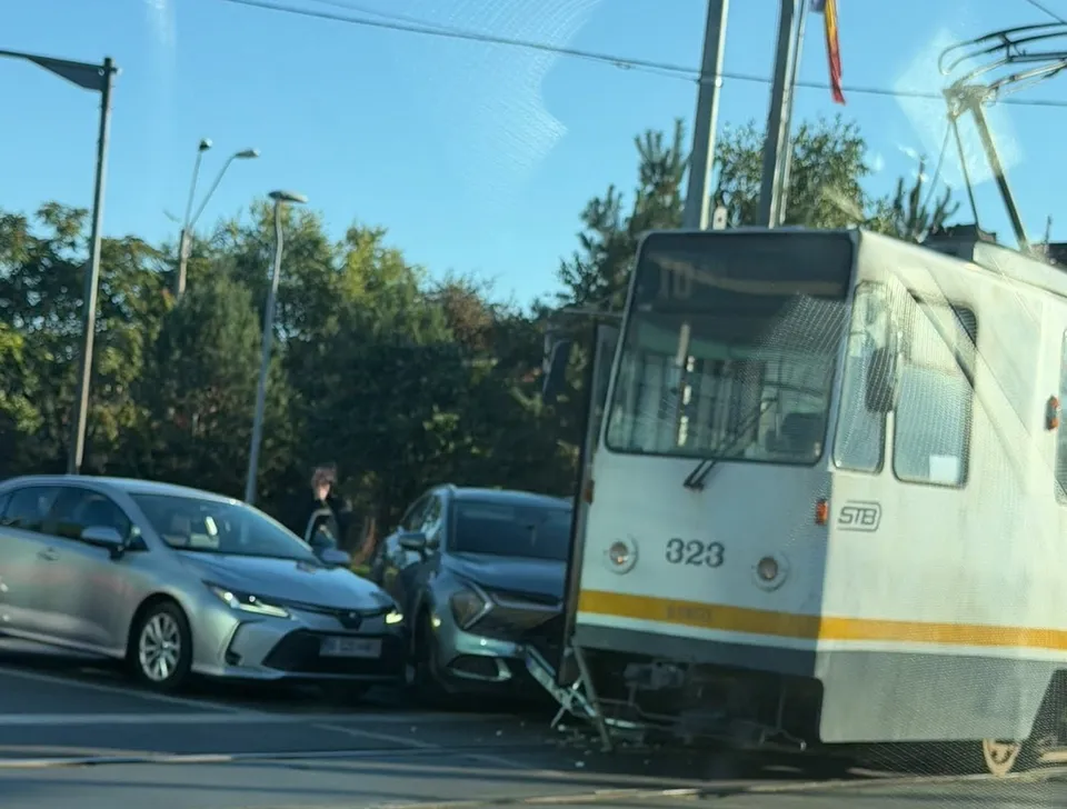 Un tramvai alb cu numărul 323 este poziționat lângă două autoturisme pe o stradă cu copaci în fundal.