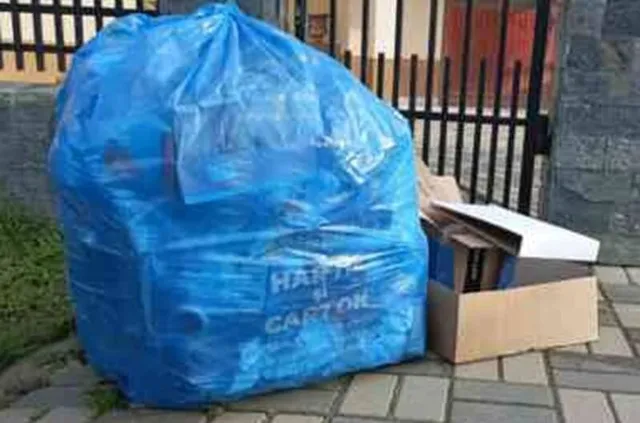 Un sac mare de plastic albastru umplut cu obiecte este așezat pe o trotuar lângă o cutie de carton deschisă.