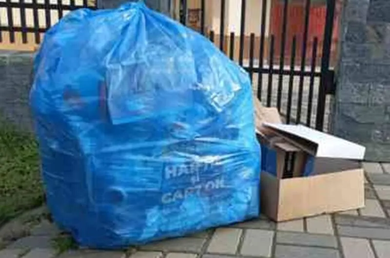 Un sac mare de plastic albastru umplut cu obiecte este așezat pe o trotuar lângă o cutie de carton deschisă.