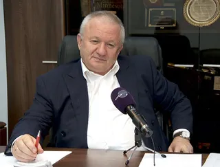 Un bărbat cu sacou închis și cămașă albă stă la o masă cu un microfon în față și un obiect roșu în mână.