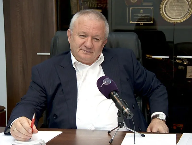 Un bărbat cu sacou închis și cămașă albă stă la o masă cu un microfon în față și un obiect roșu în mână.