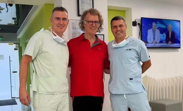 Trei bărbați stau în picioare într-o încăpere, doi purtând uniforme medicale deschise la gât și unul purtând un tricou roșu.