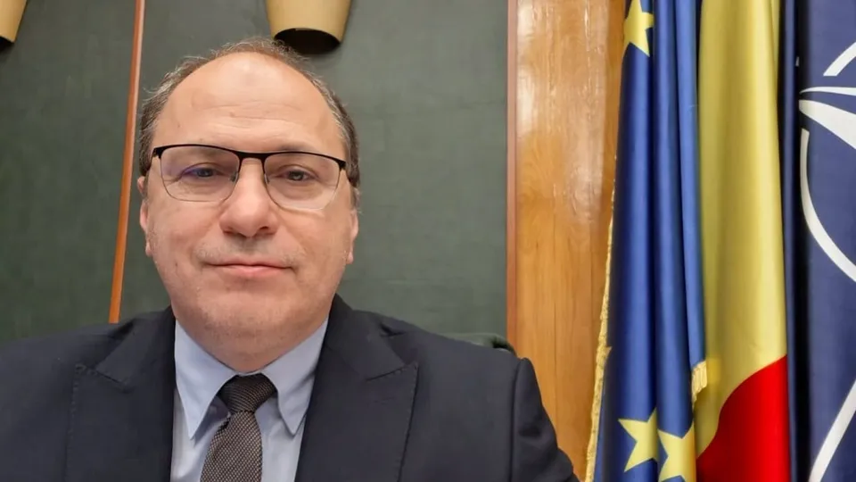Un bărbat în costum de afaceri cu cravată, ochelari și păr încălțit, privind direct camera, în fața unui fond verde cu steagul UE, românesc și NATO.