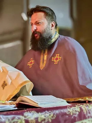 Un bărbat cu barbă lungă poartă o robă purpurie decorată cu cruci aurii, stând lângă o masă cu un volum deschis.