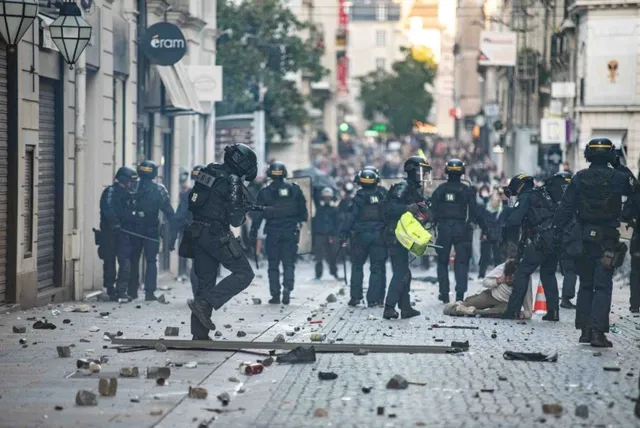 O grupă de polițiști în echipament de protecție întunecat stă pe o stradă pavată presărată cu resturi și dezhidratează o persoană întinsă pe sol.