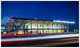O clădire modernă cu fațadă de sticlă iluminată este situată sub un cer nocturn, cu urme de lumină de la vehicule în prim-plan.
