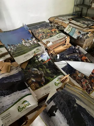 Pachete și cutii cu albume turistice, multe cu imaginea unor peisaje montane și logo-ul România, sunt aruncate și depozitate haotic pe podea și pe rafturi de metal.