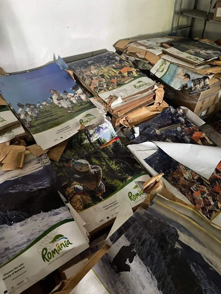 Pachete și cutii cu albume turistice, multe cu imaginea unor peisaje montane și logo-ul România, sunt aruncate și depozitate haotic pe podea și pe rafturi de metal.