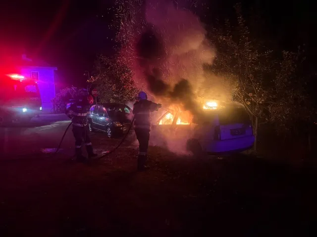 Două persoane în uniforme înt dark folosesc un furtun pentru a stinge un incendiu care afectează un autoturism parcat noaptea.
