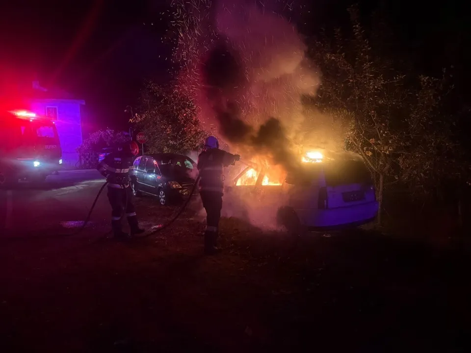 Două persoane în uniforme înt dark folosesc un furtun pentru a stinge un incendiu care afectează un autoturism parcat noaptea.