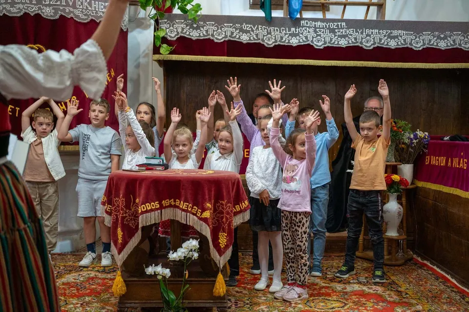 Un grup de copii stă în picioare în fața unei mese acoperite cu un drapel roșu, ridicând mâinile în sus într-o încăpere interioară.