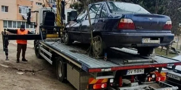 Un autovehicul de culoare închisă este încărcat pe platforma unui camion de remorcare aflat lângă un operator îmbrăcat în vestă portocalie.