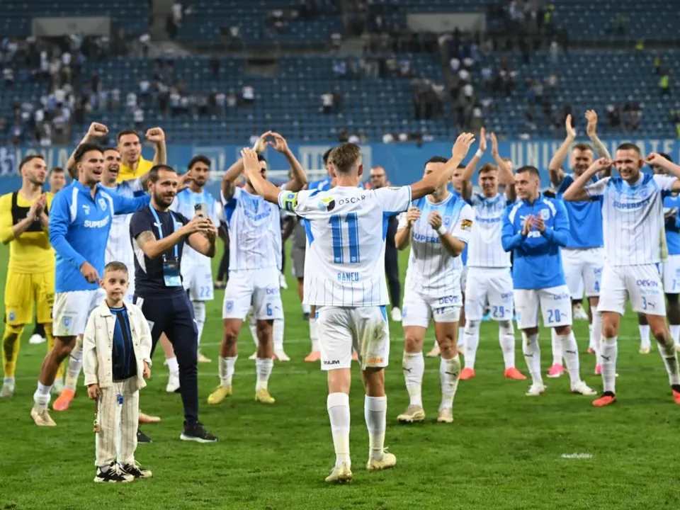 Un grup de fotbaliști în echipamente albastre și albe sărbătoresc pe un teren de fotbal în fața unor tribune cu spectatori.