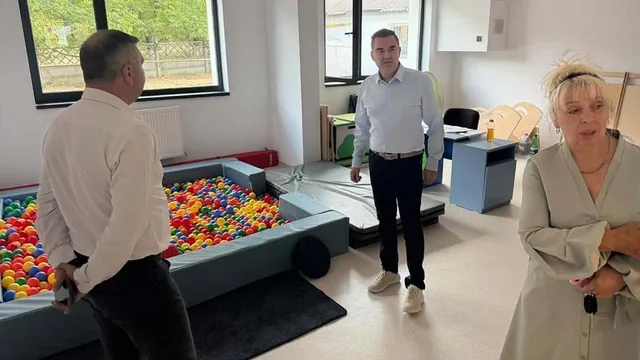 Trei persoane stau într-o încăpere cu o piscină de mingi colorate, o saltea de gimnastică și mobilier de copii.