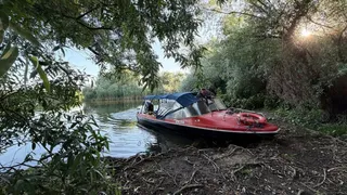 O barcă roșie cu acoperiș textil albastru este parcată pe malul unui râu lângă vegetație densă și copaci sub lumina soarelui.