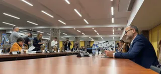 Un grup de persoane stă la o masă lungă de conferințe într-o încăpere iluminată de corpuri de iluminat lungi și albe.