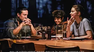 Three people are sitting at a dining table with glasses, a thermos, and plates, engaged in conversation.