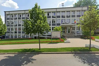 O clădire administrativă cu ferestre multiple este înconjurată de iarbă verde, flori roșii și copaci sub un cer cu nori albi.