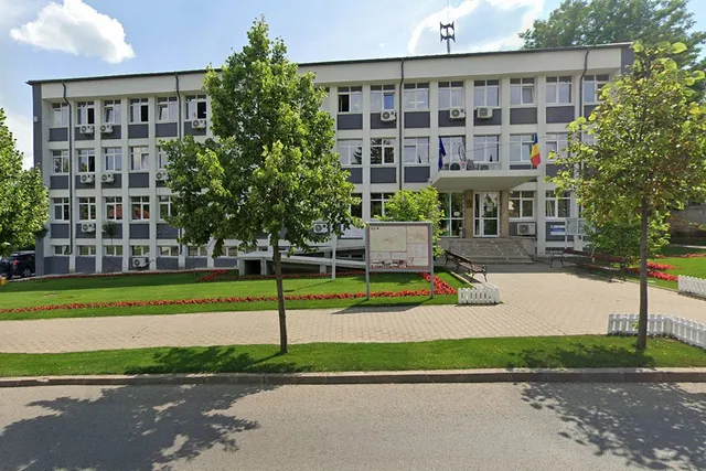 O clădire administrativă cu ferestre multiple este înconjurată de iarbă verde, flori roșii și copaci sub un cer cu nori albi.
