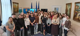 Un grup de tineri stă în picioare într-o încăpere cu tablouri pe pereți și ferestre cu perdele albe.