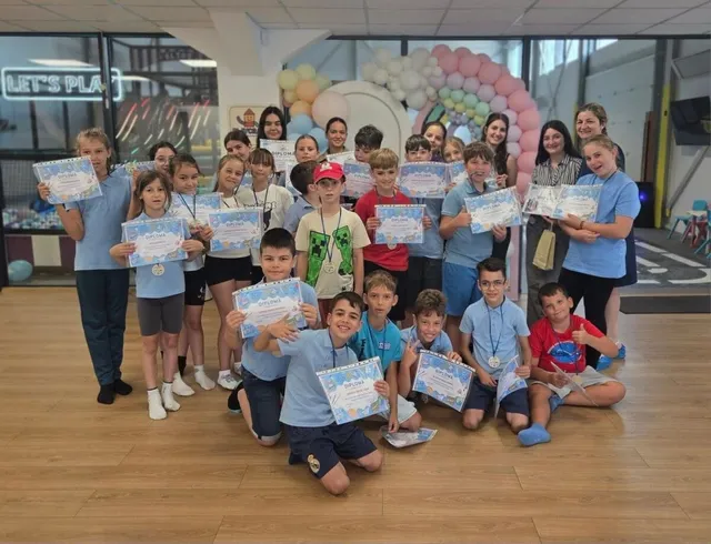 Un grup de copii și adolescenți stau în grup pe un podea de lemn, ținând foi de hârtie albe cu text și grafică colorată.