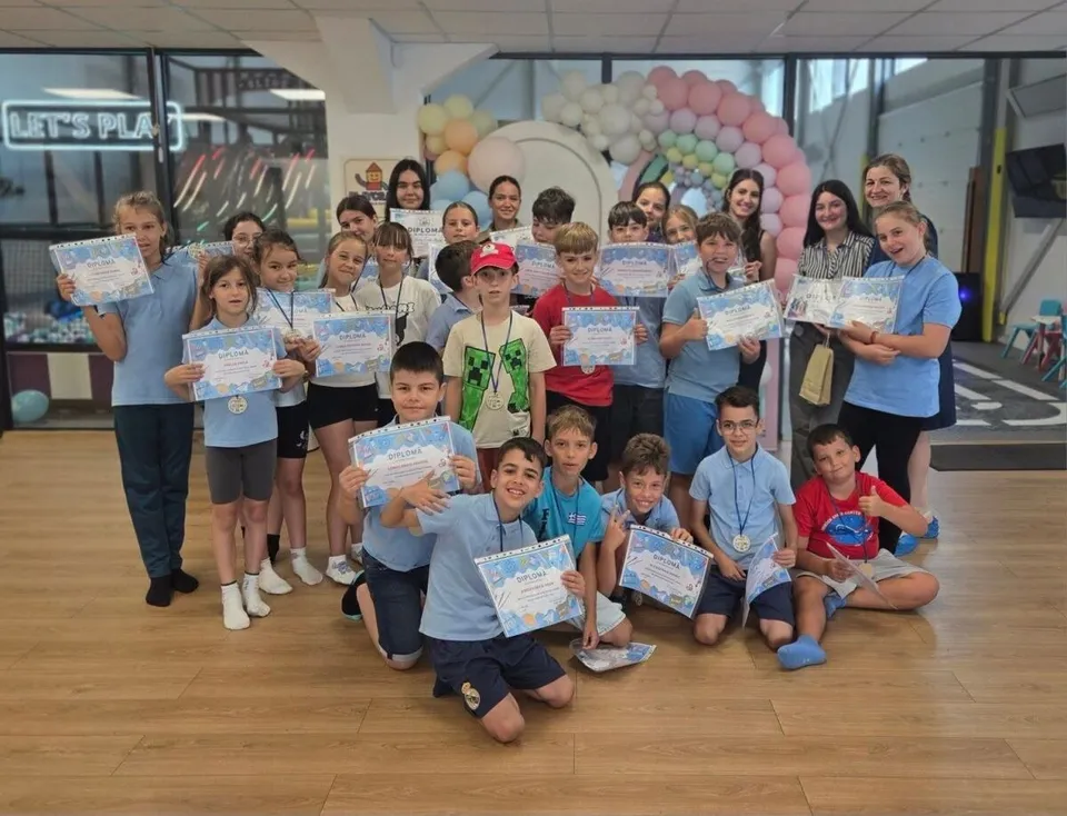 Un grup de copii și adolescenți stau în grup pe un podea de lemn, ținând foi de hârtie albe cu text și grafică colorată.