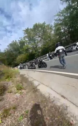 O persoană cu blugi și o jachetă de culoare închisă stă lângă mai multe motociclete parcate pe marginea unui drum asfaltat lângă un gard metalic.